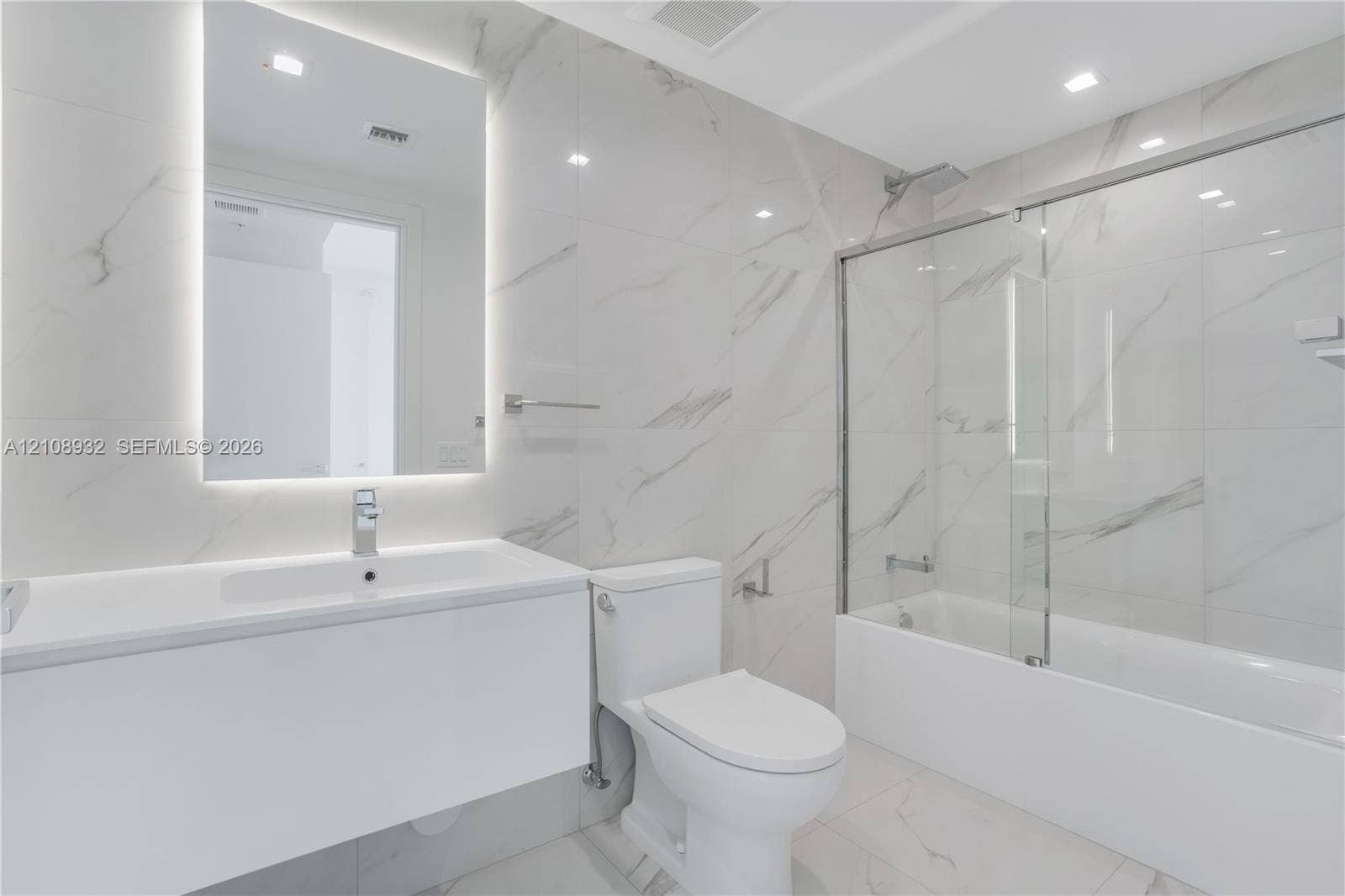 1170 E Hallandale Beach Boulevard, #1201, Hallandale Beach, FL 33009 Bathroom featuring large-format stone-patterned wall and floor tile, a modern floating vanity with an integrated sink, a frameless glass tub enclosure, and an LED-backlit mirror
