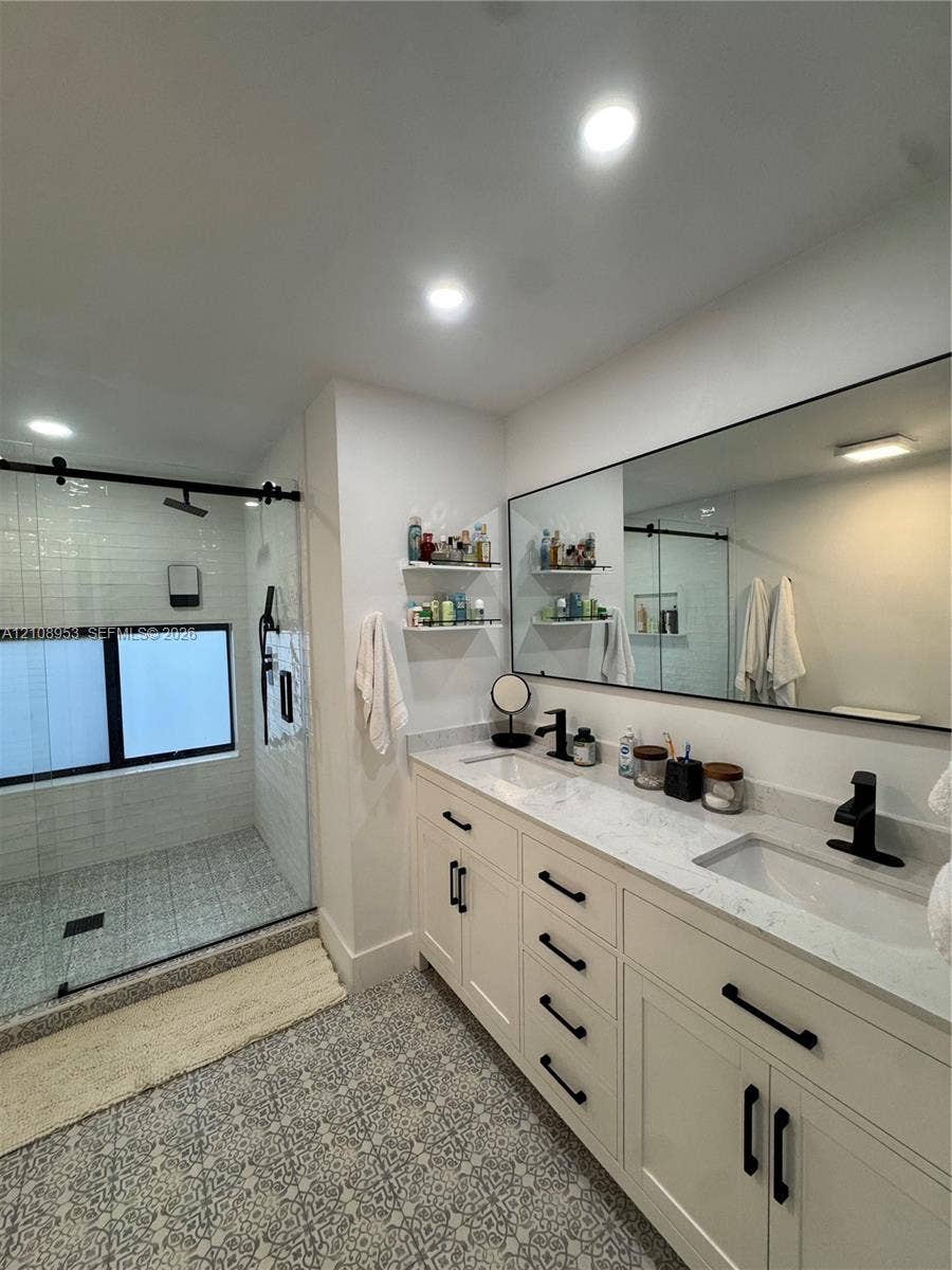 Double vanity with stone-look countertops, matte black fixtures, walk-in shower with glass enclosure, and patterned tile flooring