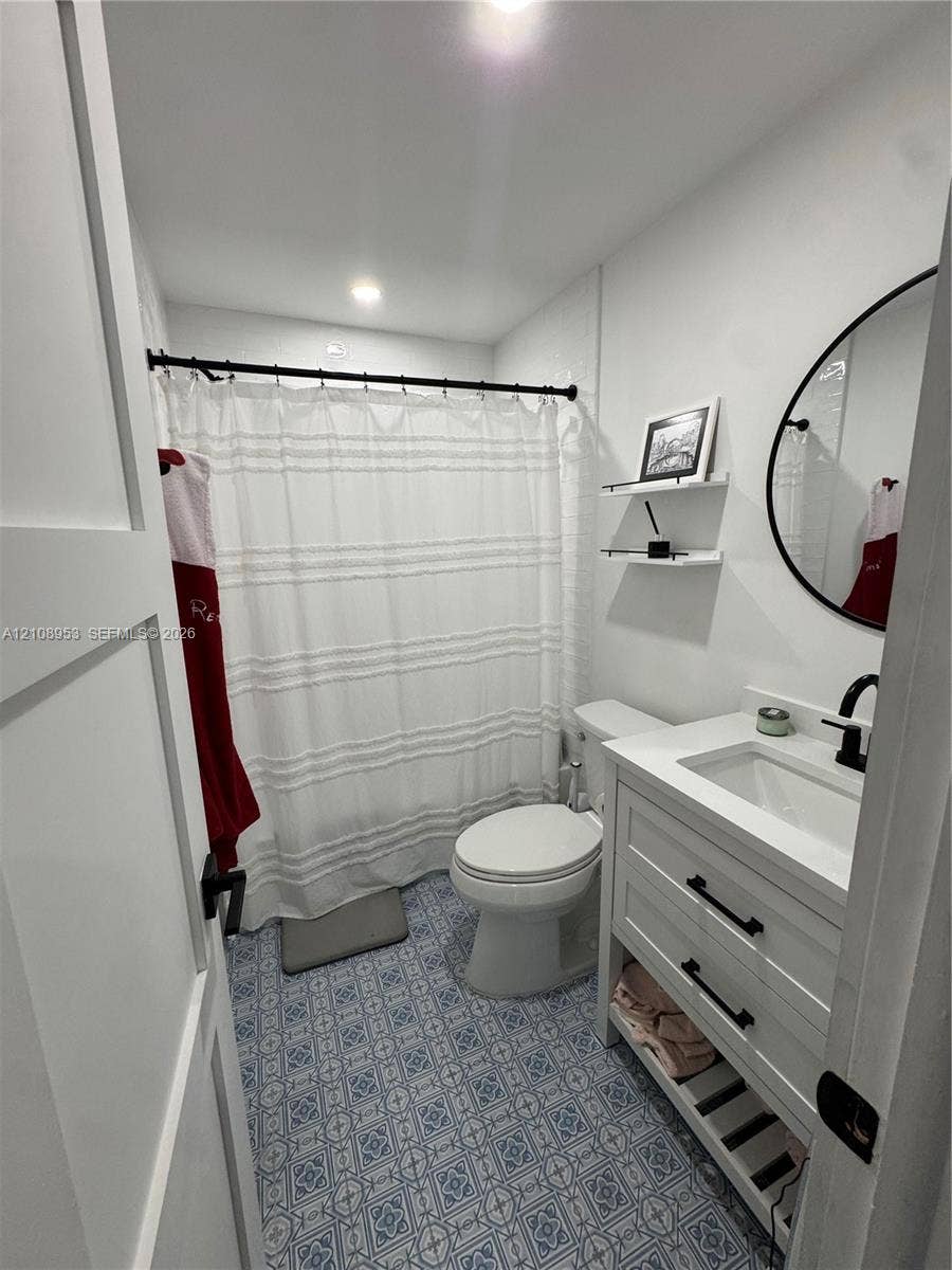 Bathroom featuring patterned tile flooring, white vanity with integrated sink and matte black hardware, round vanity mirror, recessed ceiling lighting, and wall-mounted shelving