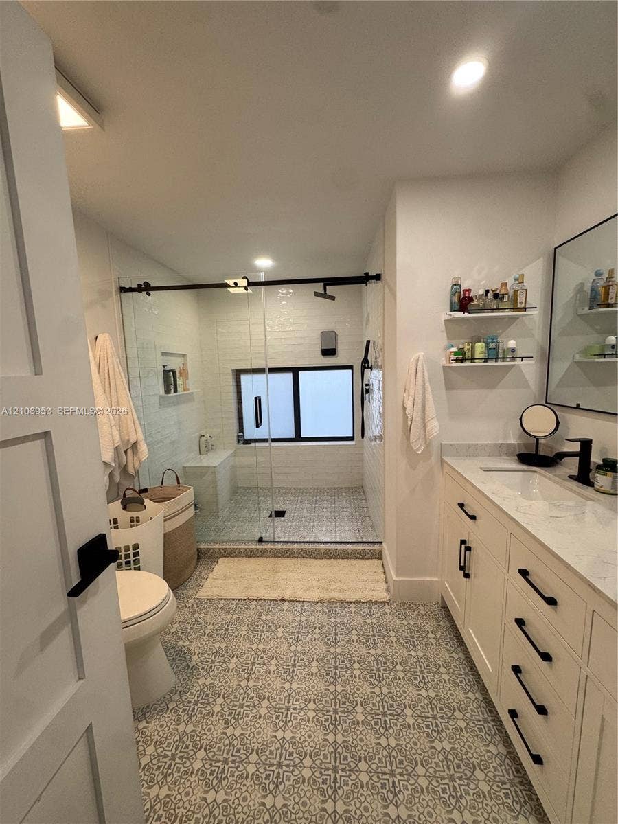 Custom cabinetry with dark hardware, stone-top vanity, walk-in glass-enclosed shower with matte black fixtures, patterned tile flooring, and integrated wall shelving