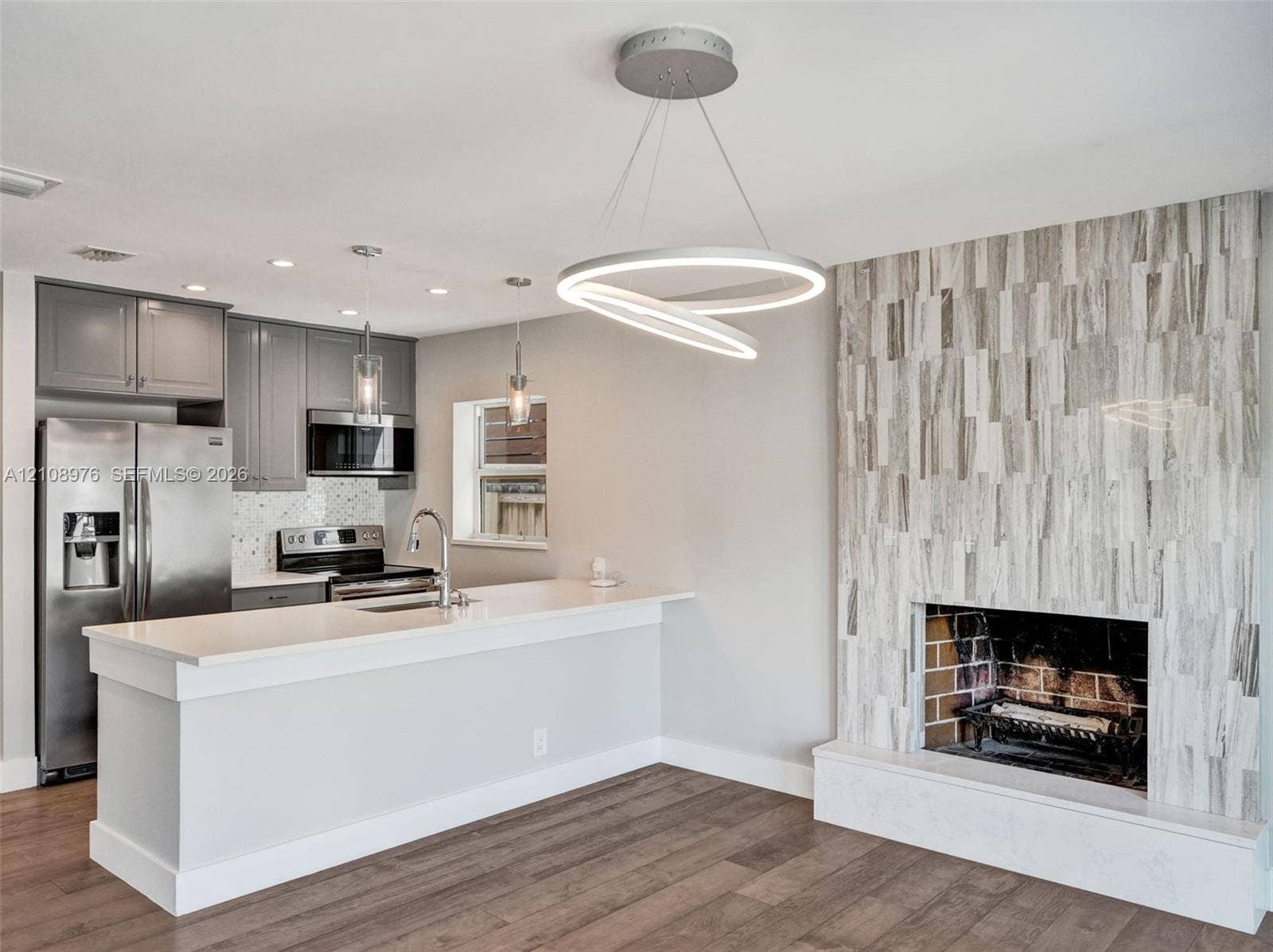 525 SW 12th Street, Fort Lauderdale, Fort Lauderdale, FL 33315 Kitchen featuring grey cabinetry, stainless steel appliances, and a peninsula with a quartz countertop, complemented by a floor-to-ceiling fireplace with stone veneer surround and wood-finish flooring
