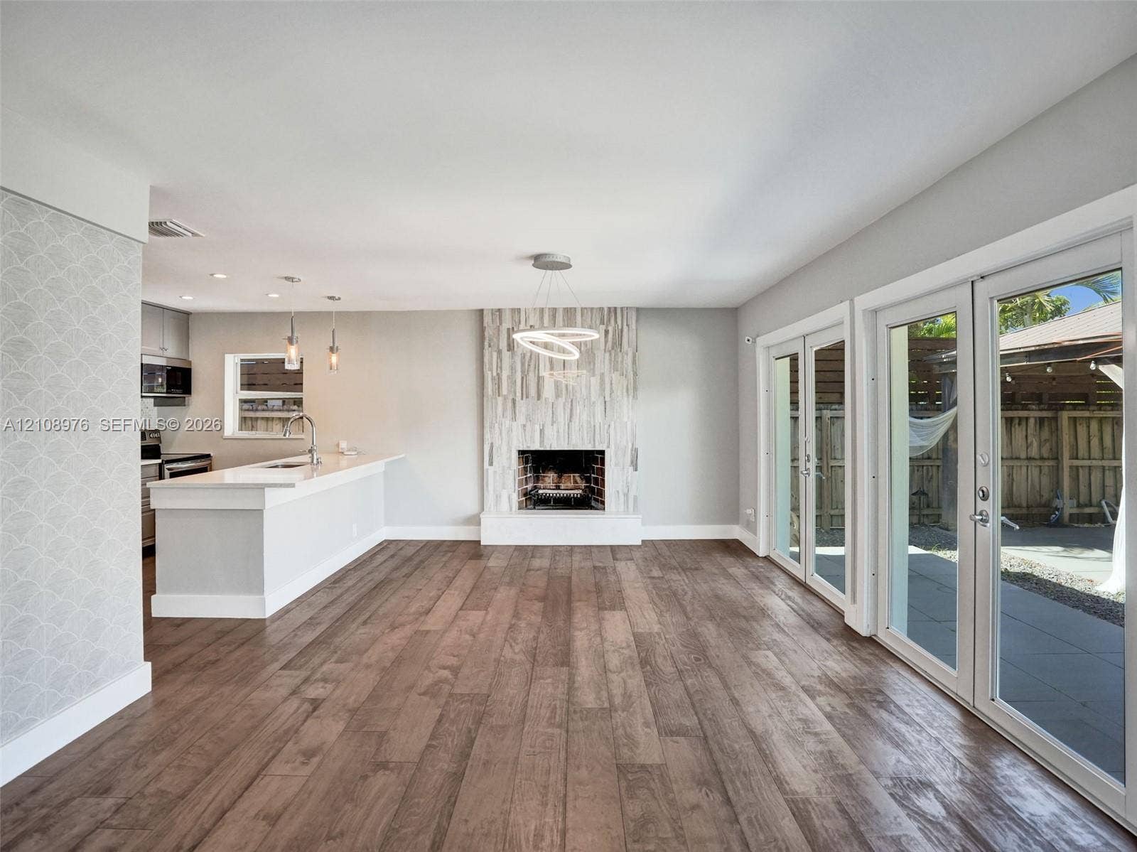 525 SW 12th Street, Fort Lauderdale, Fort Lauderdale, FL 33315 Open-concept interior featuring wood-finish flooring, floor-to-ceiling fireplace with vertical stone-finish surround, kitchen island with solid-surface countertop, dual glass-paned doors, and contemporary tiered chandelier