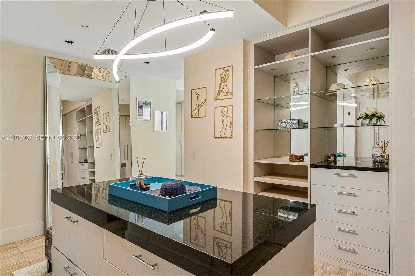Custom walk-in closet featuring floor-to-ceiling cabinetry, a central storage island with a dark stone countertop, built-in glass shelving, mirrored paneling, and a contemporary curved light fixture