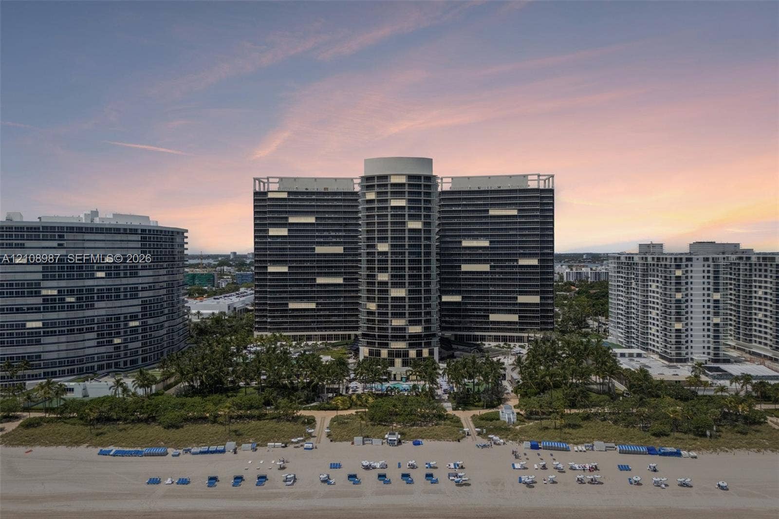 High-rise beachfront structure featuring wraparound balconies, expansive glass facade, landscaped grounds with mature palm trees, and direct sand access