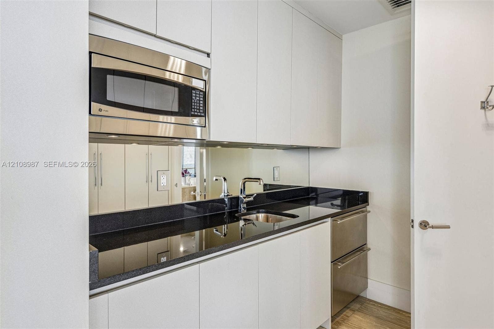 Kitchenette featuring white flat-panel cabinetry, dark stone countertops, mirrored backsplash, stainless steel microwave, and stainless steel under-counter drawer refrigerator