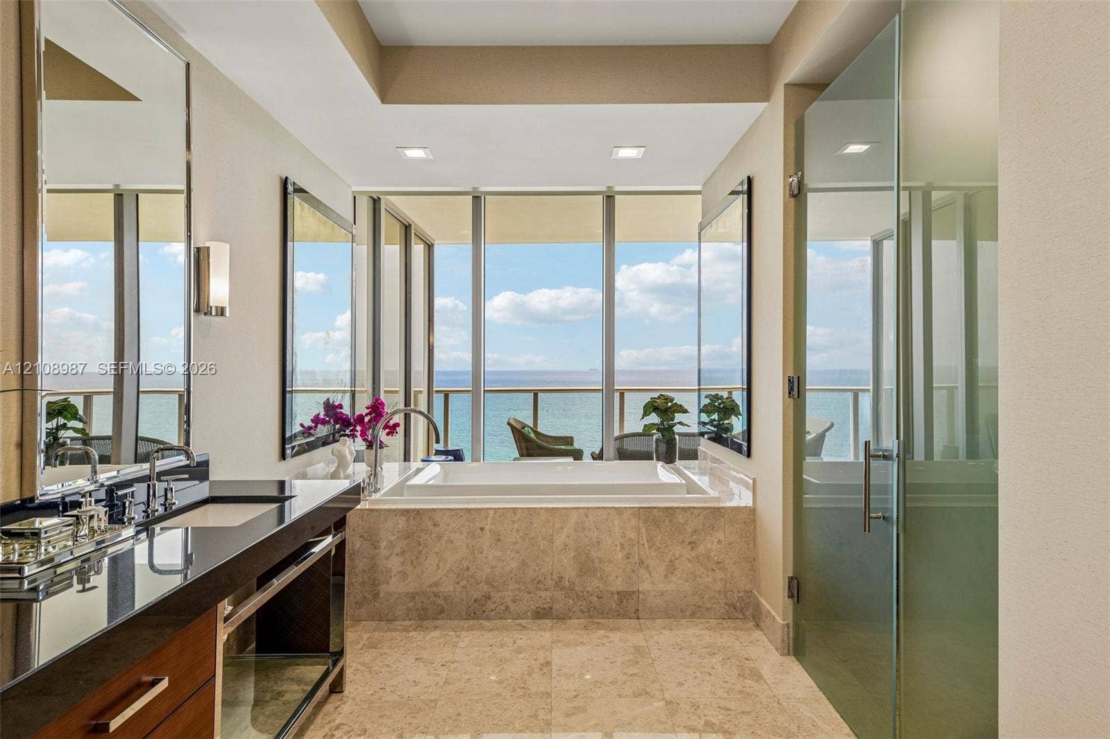 Bathroom featuring a stone-encased soaking tub, floor-to-ceiling windows, dark stone-surfaced vanity with dual sinks, and a glass-enclosed shower
