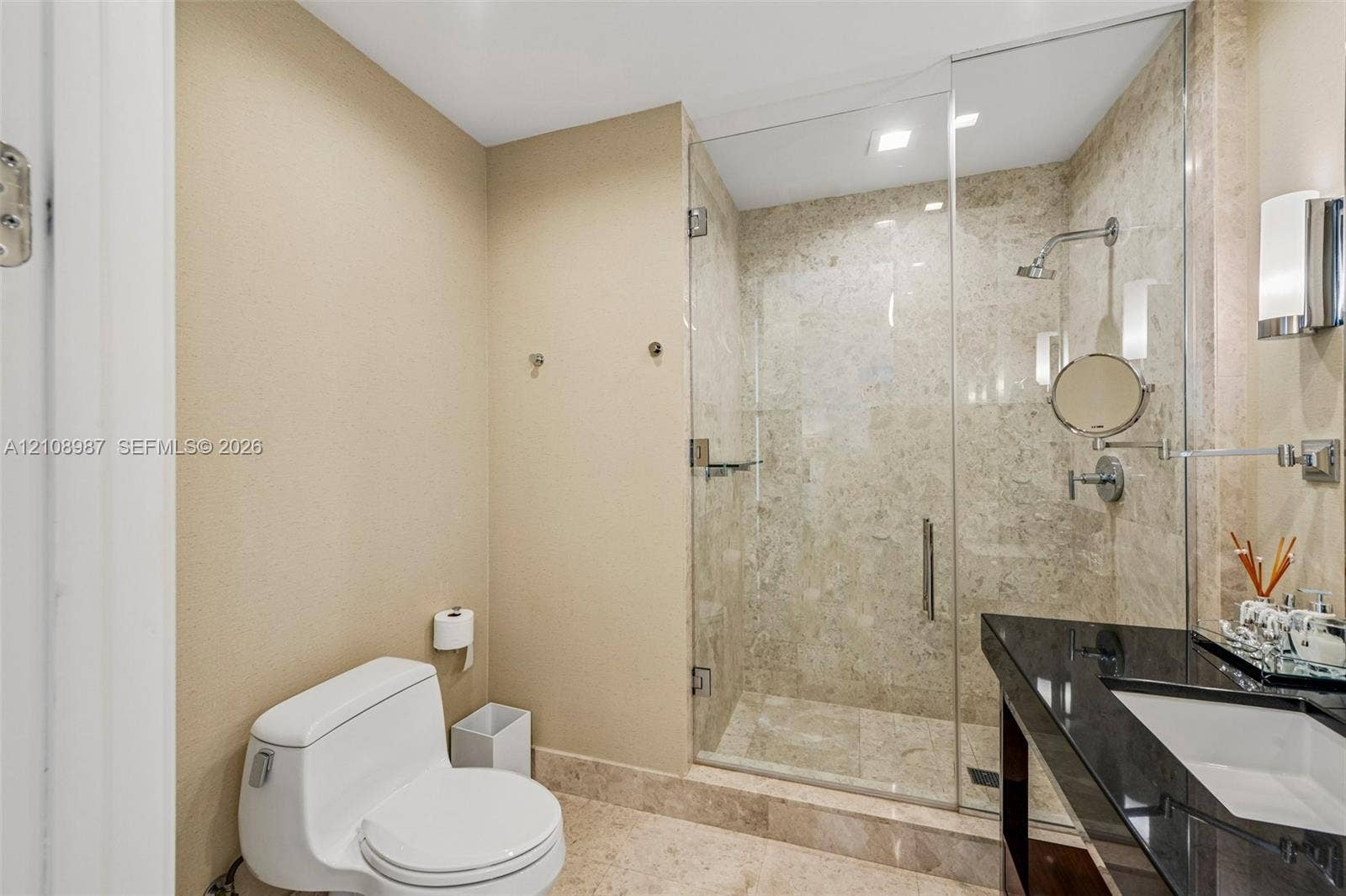 Frameless glass-enclosed shower with stone-tile surround, vanity featuring a polished dark stone countertop with an integrated basin, and stone flooring