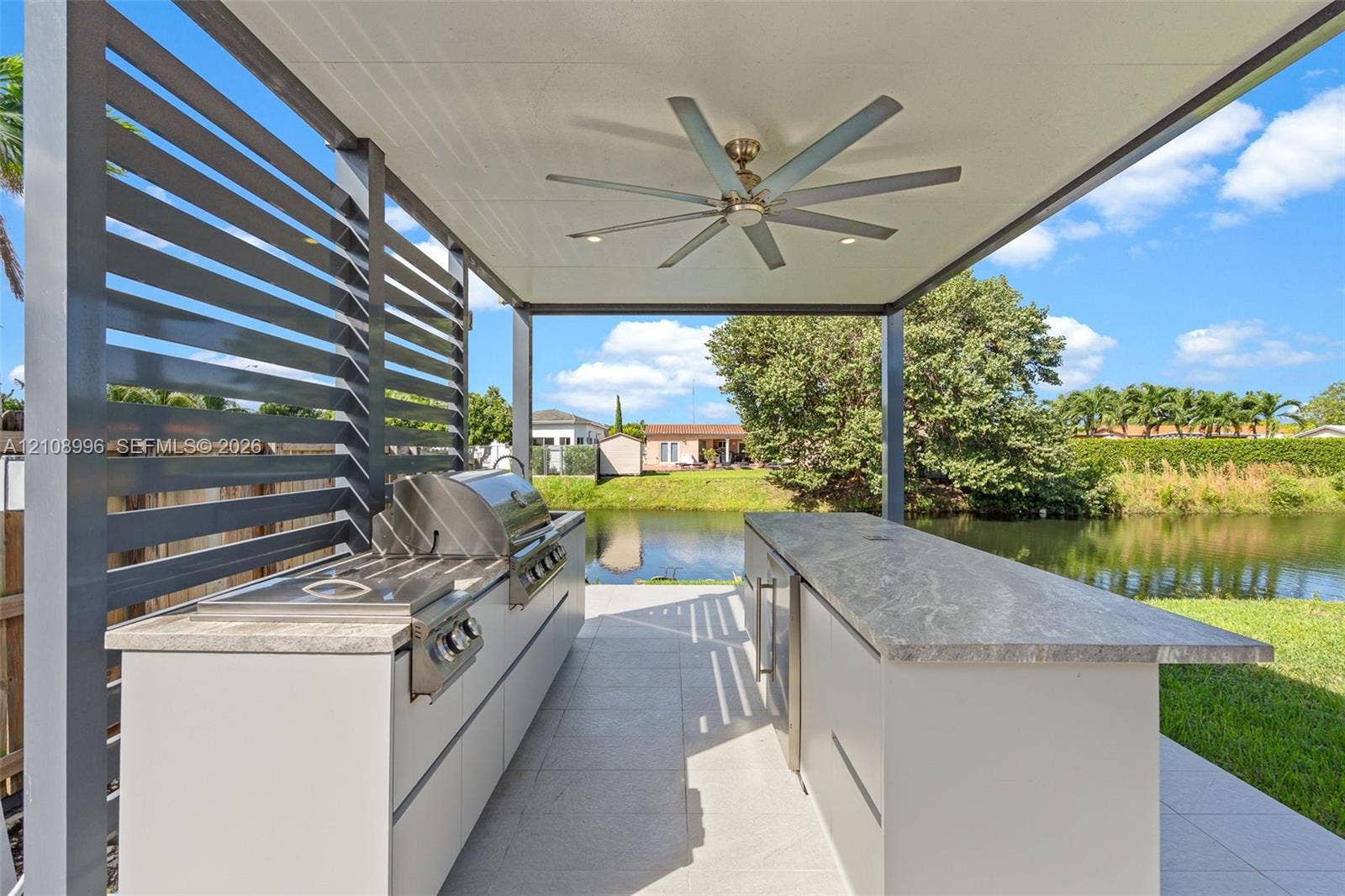 4252 SW 131st Avenue, Miami, FL 33175 Covered outdoor kitchen featuring a stone-topped island, stainless steel grill, horizontal slat privacy screen, and an integrated ceiling fan