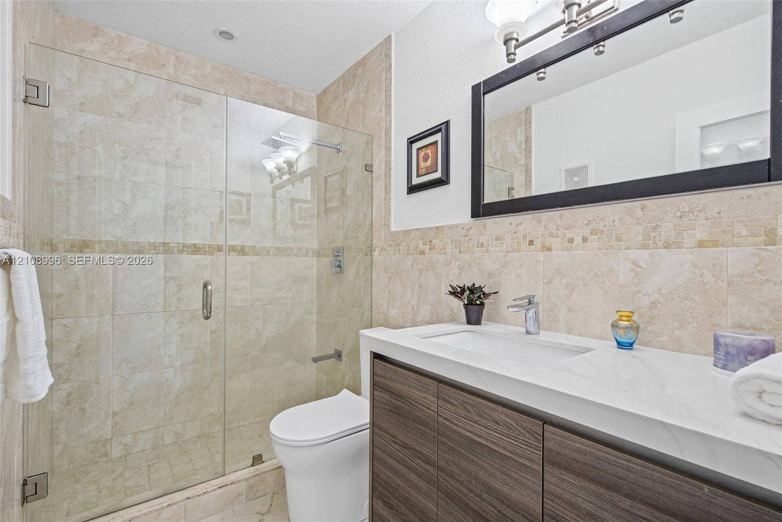 4252 SW 131st Avenue, Miami, FL 33175 Bathroom featuring a vanity with wood-finish cabinetry, a white countertop with an integrated sink, a frameless glass-enclosed shower with neutral tile surround, and a metal-framed mirror