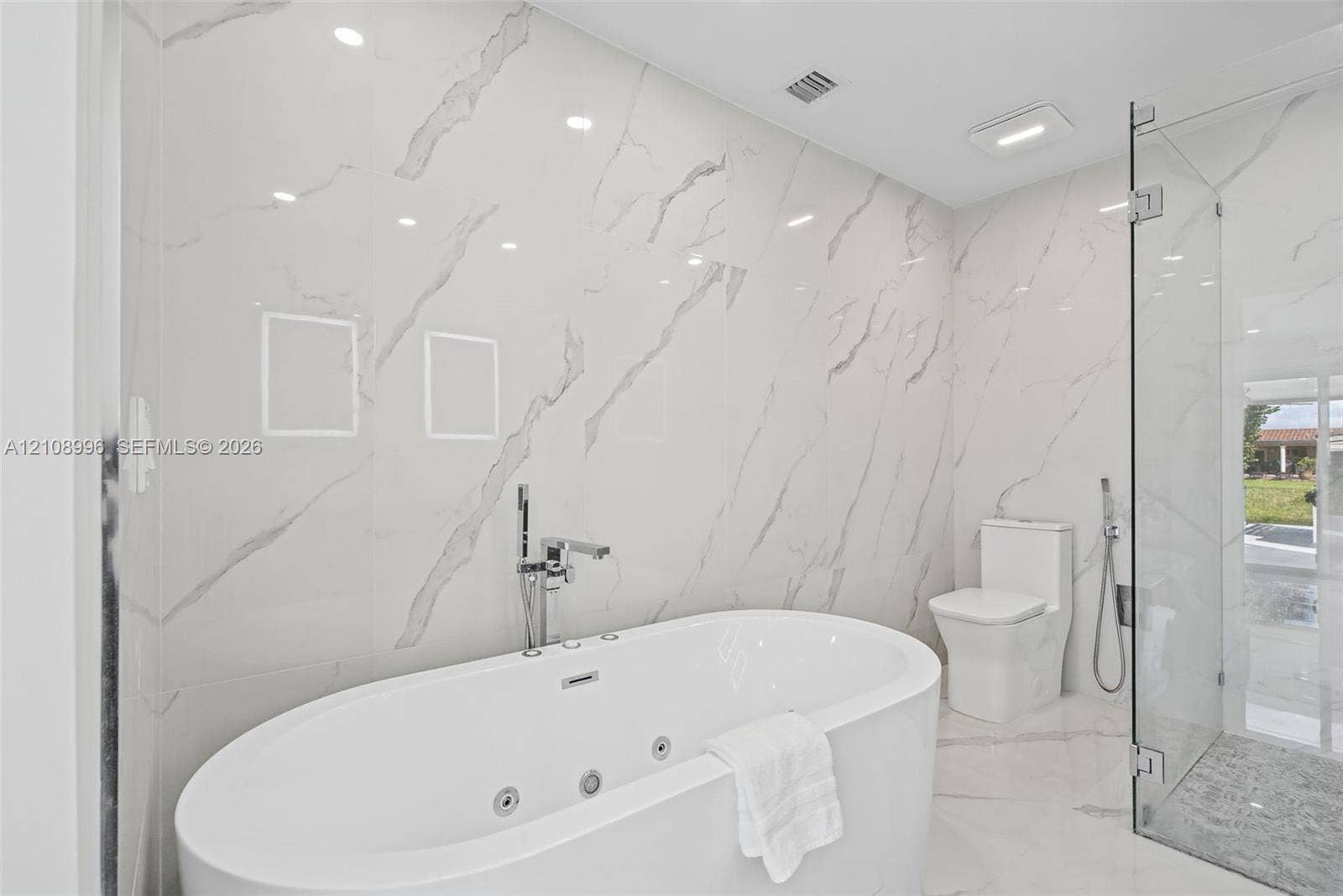 4252 SW 131st Avenue, Miami, FL 33175 Freestanding jetted soaking tub with deck-mounted chrome faucet, floor-to-ceiling large-format stone-finish tile wall, frameless glass enclosed shower, and white dual-flush toilet