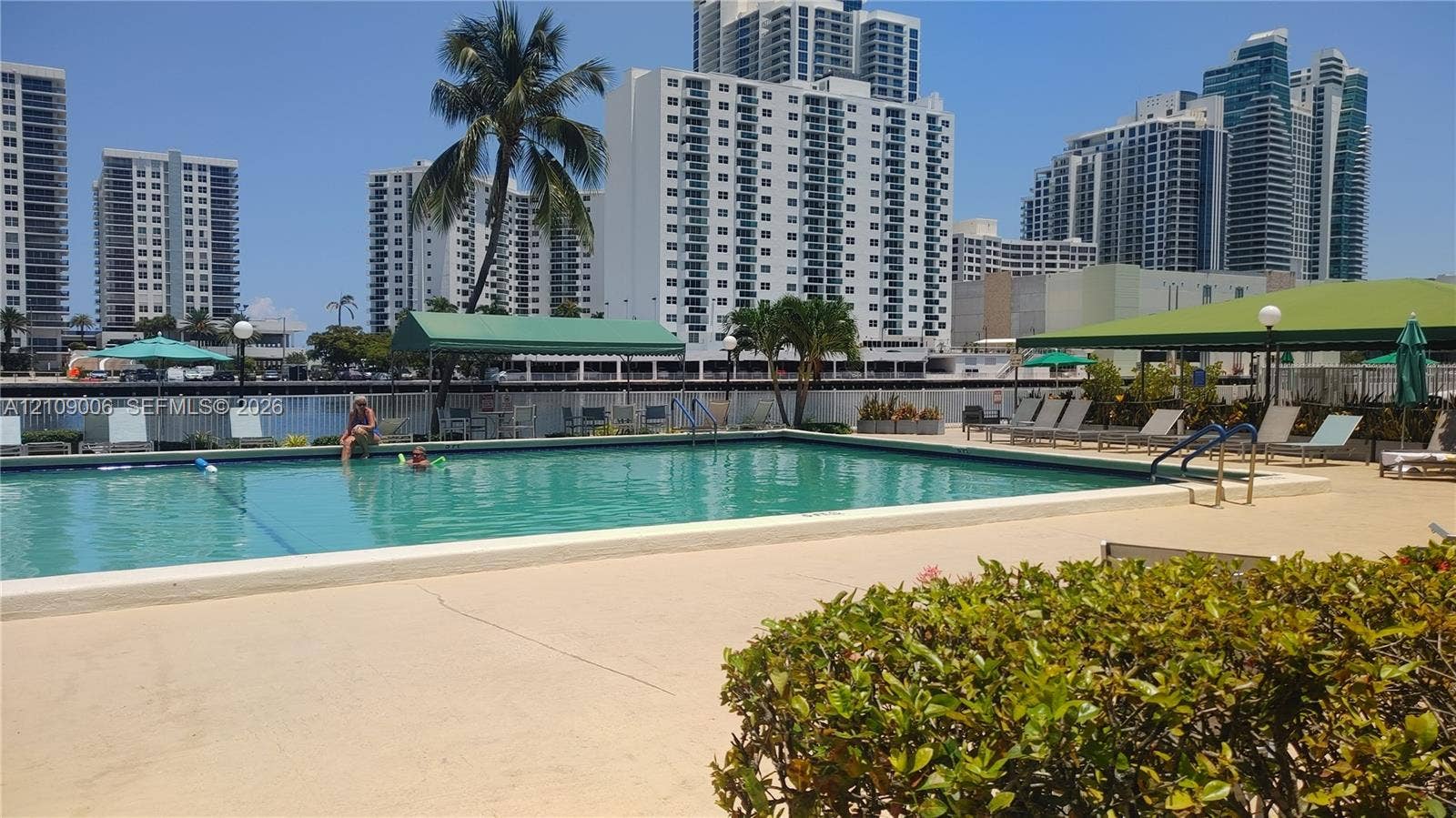 In-ground swimming pool featuring a concrete deck, perimeter fencing, overhead canopy structures, and mature landscaping