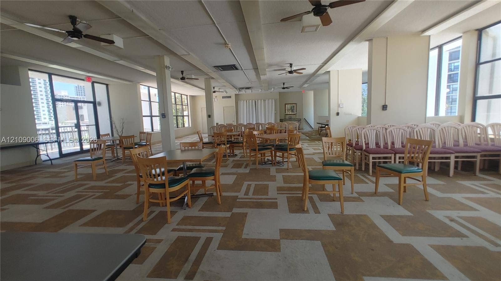 Expansive interior space featuring patterned carpeting, multiple ceiling fans, floor-to-ceiling window panels, and neutral wall tones