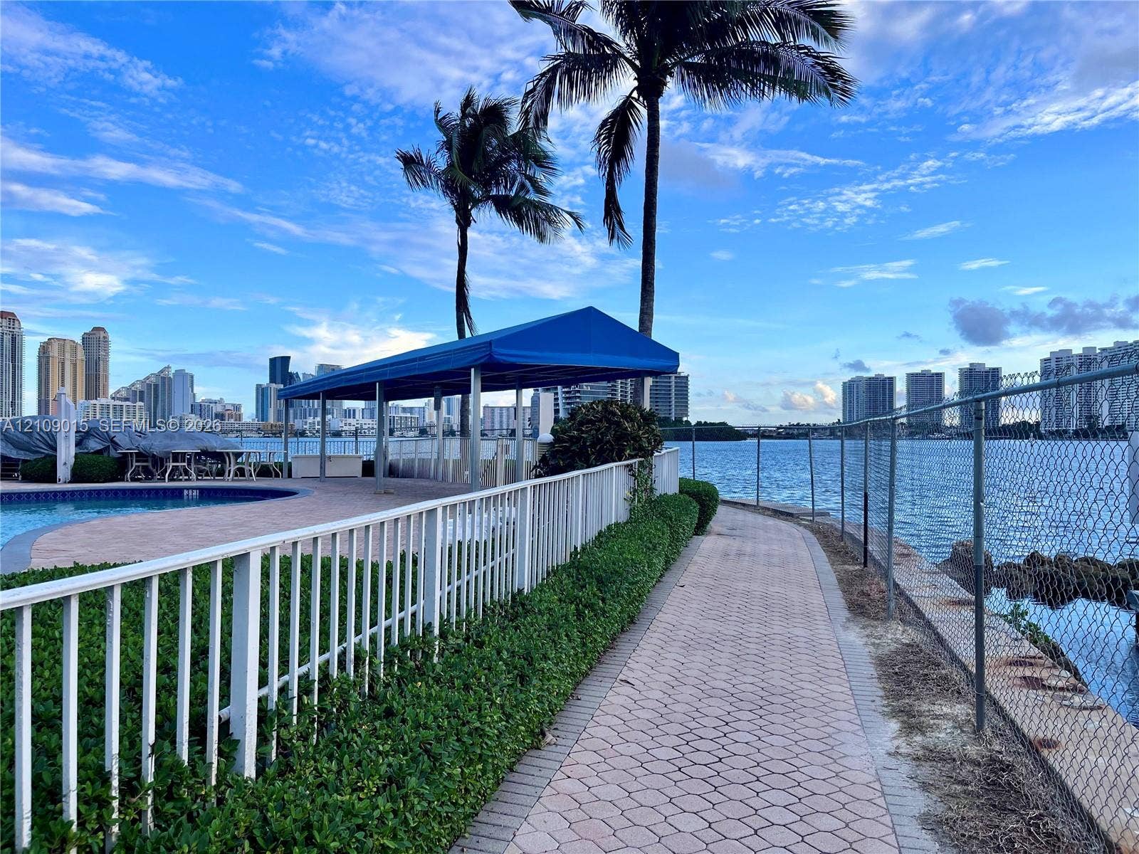 3530 Mystic Pointe Drive, #1709, Aventura, FL 33180 Waterfront paver walkway featuring metal perimeter fencing, mature palm trees, a poolside canopy structure, and proximity to the canal