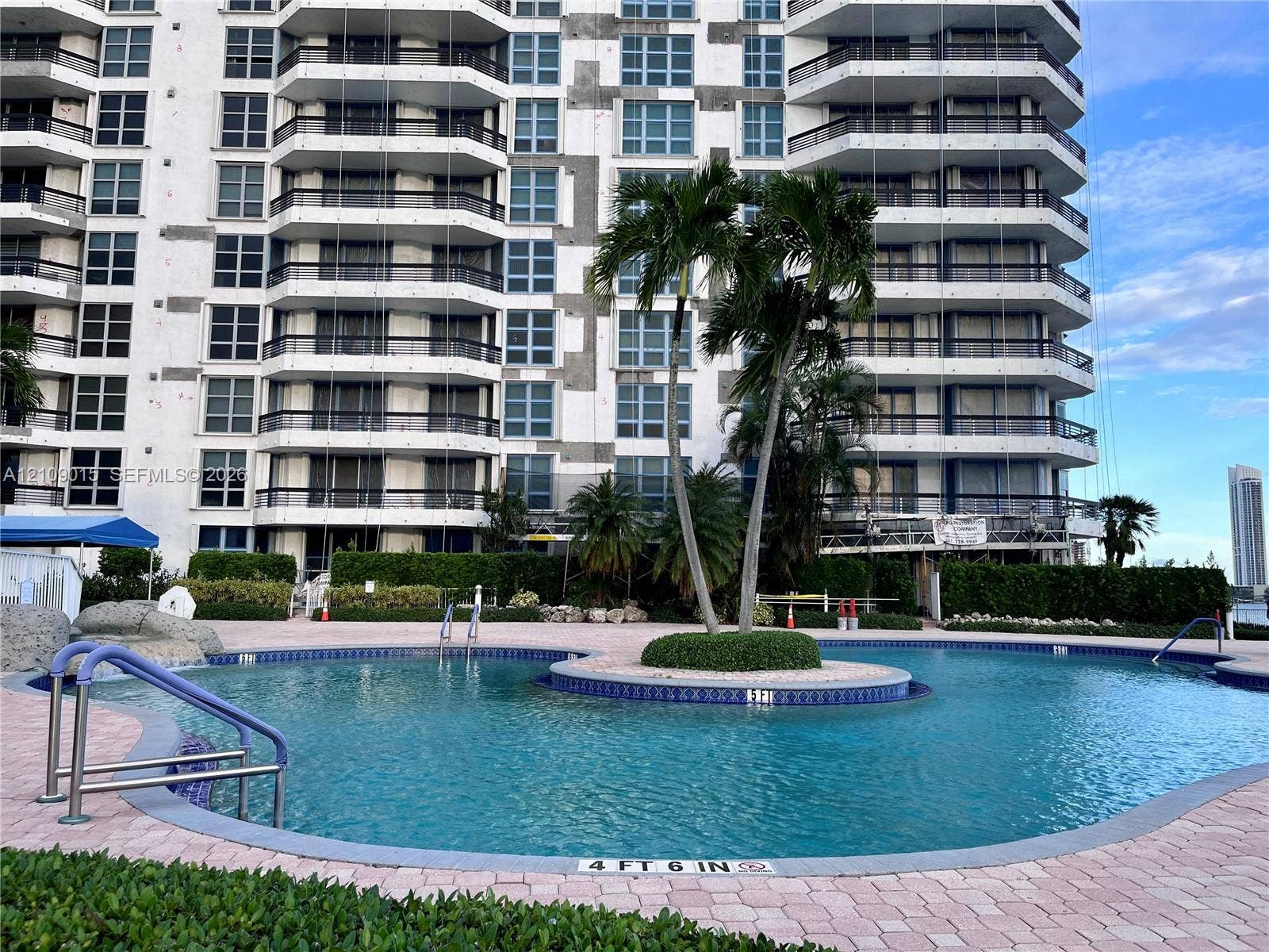 3530 Mystic Pointe Drive, #1709, Aventura, FL 33180 Curved swimming pool featuring tiled coping, metal handrails, and a central landscaped island with palm trees, set within a paved patio area