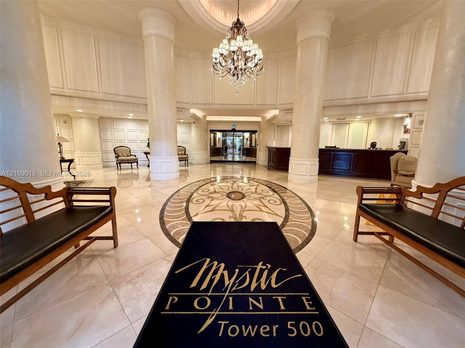 3530 Mystic Pointe Drive, #1709, Aventura, FL 33180 Grand lobby featuring high ceilings with architectural columns, stone tile flooring with decorative medallion inlay, and an ornate crystal chandelier