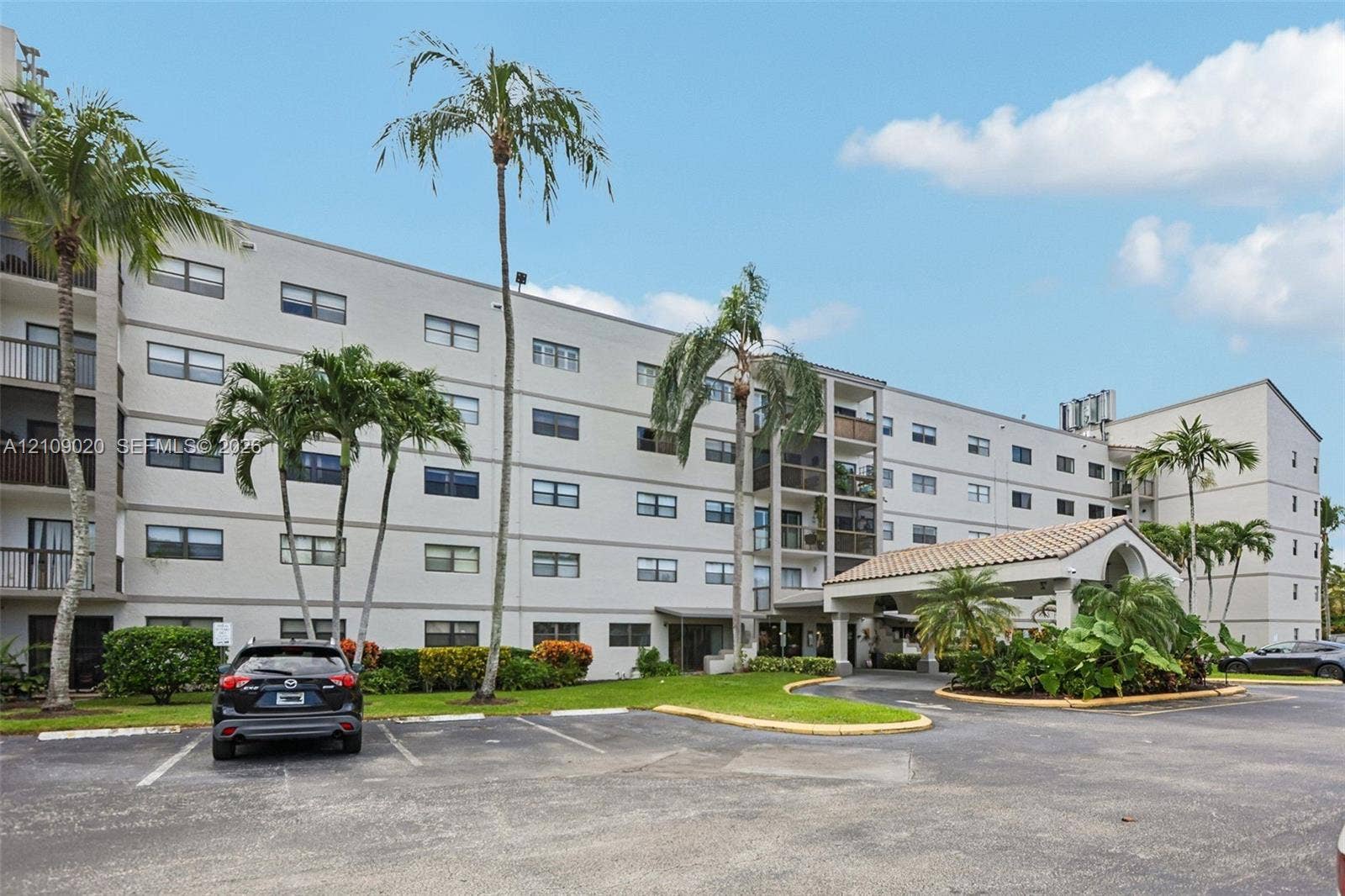 701 NW 19th Street, #300, Fort Lauderdale, FL 33311 Mid-rise residential building exterior featuring a covered entryway with a tile roof, paved surface parking, and mature palm landscaping