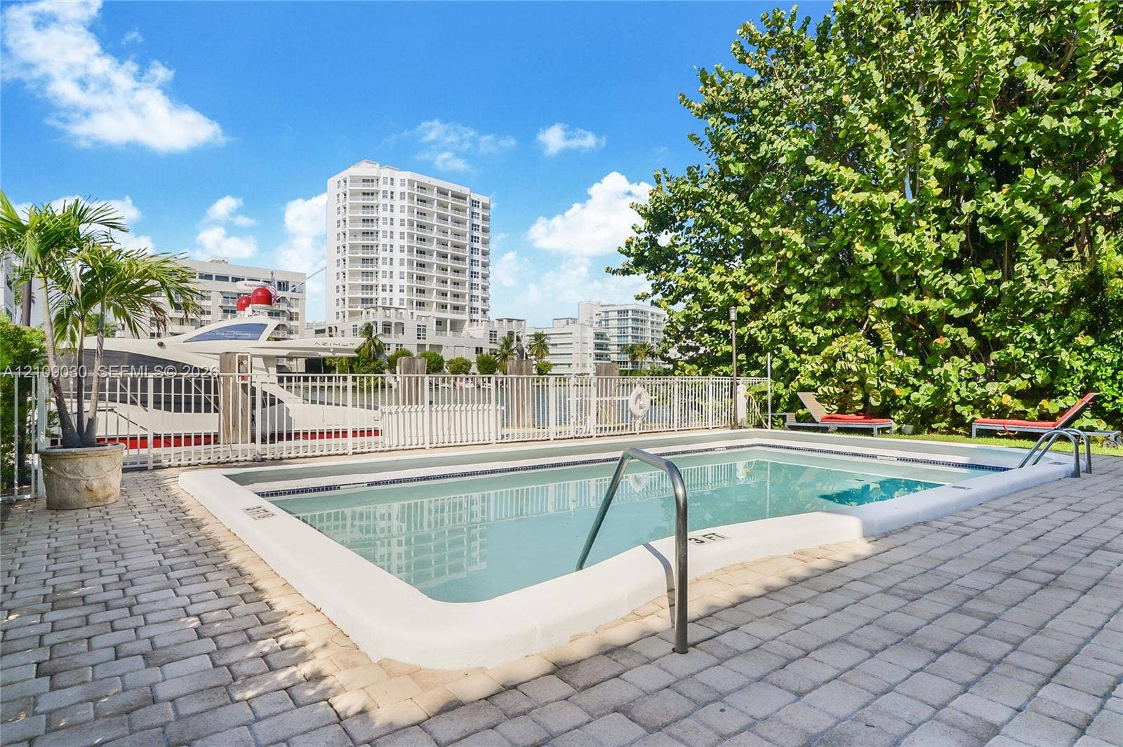 Rectangular inground swimming pool featuring a light-toned coping, metal handrails, and a surrounding stone-paver deck, bordered by a white metal perimeter fence and mature landscaping