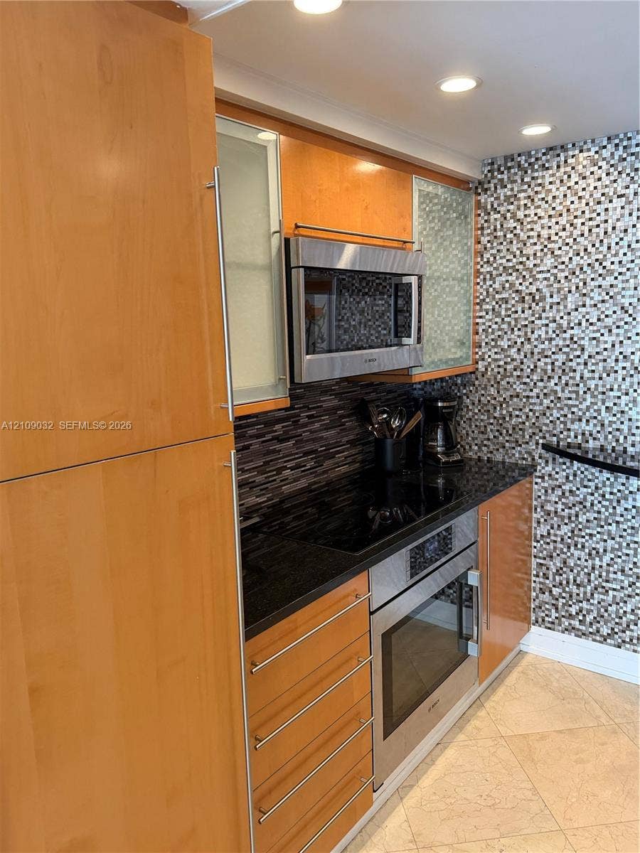 Kitchen featuring flat-panel wood cabinetry, integrated stainless steel appliances, dark stone countertops, a mosaic tile backsplash, and recessed lighting