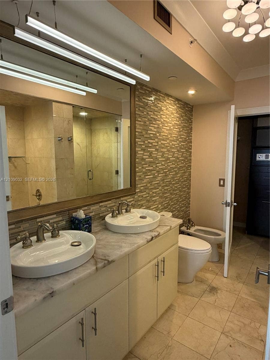 Dual vessel sinks with metal fixtures, stone countertop, mosaic tile backsplash, and oversized wall-mounted mirror