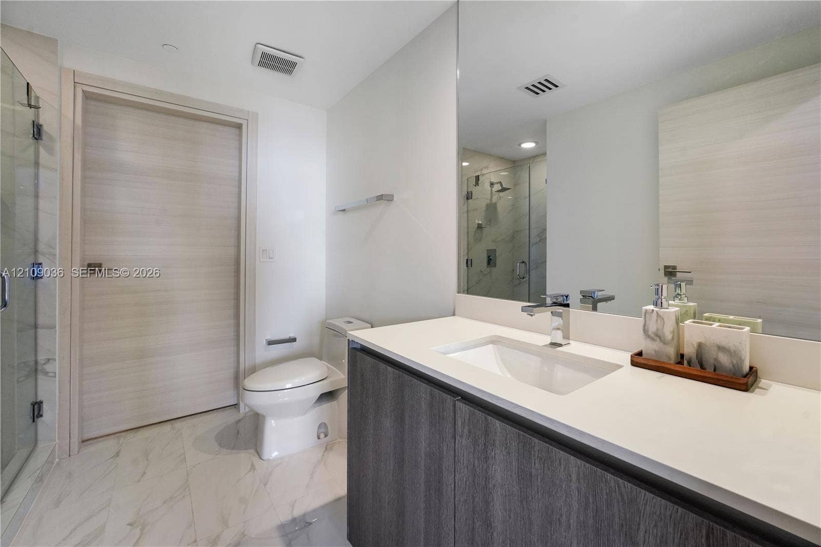 Modern vanity with a light-toned countertop, integrated basin, and dark wood-finish cabinetry, paired with large-format stone-finish floor tile, a full-width mirror, and a glass-enclosed shower