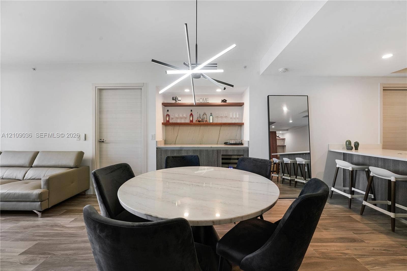 Built-in wet bar featuring stone-surface countertops, custom cabinetry, integrated wine refrigeration, and floating wood shelving, illuminated by a geometric multi-arm pendant light