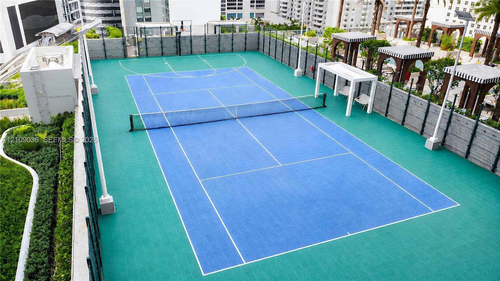 Rooftop multi-use athletic court featuring a synthetic surface, integrated basketball hoop, tennis net, and surrounding perimeter fencing with shade structures
