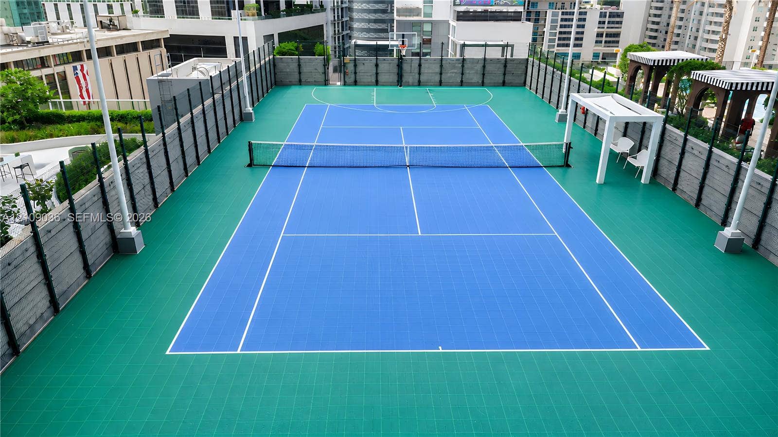 Rooftop sports court featuring a dual-purpose tennis and basketball surface, perimeter security fencing, and shaded seating structures