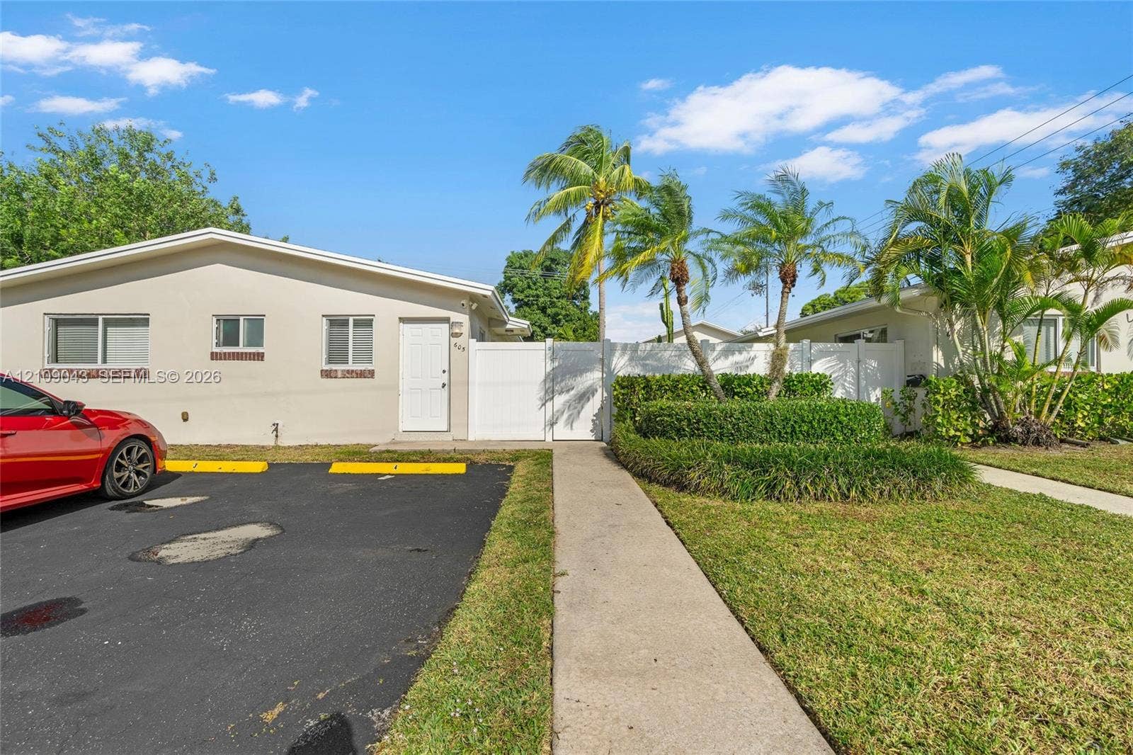 605 SW 17th Street, #1-3, Fort Lauderdale, Fort Lauderdale, FL 33315 Exterior facade featuring smooth-finish stucco, asphalt parking area, white vinyl privacy fencing, concrete walkway, and landscaped garden beds with palm trees