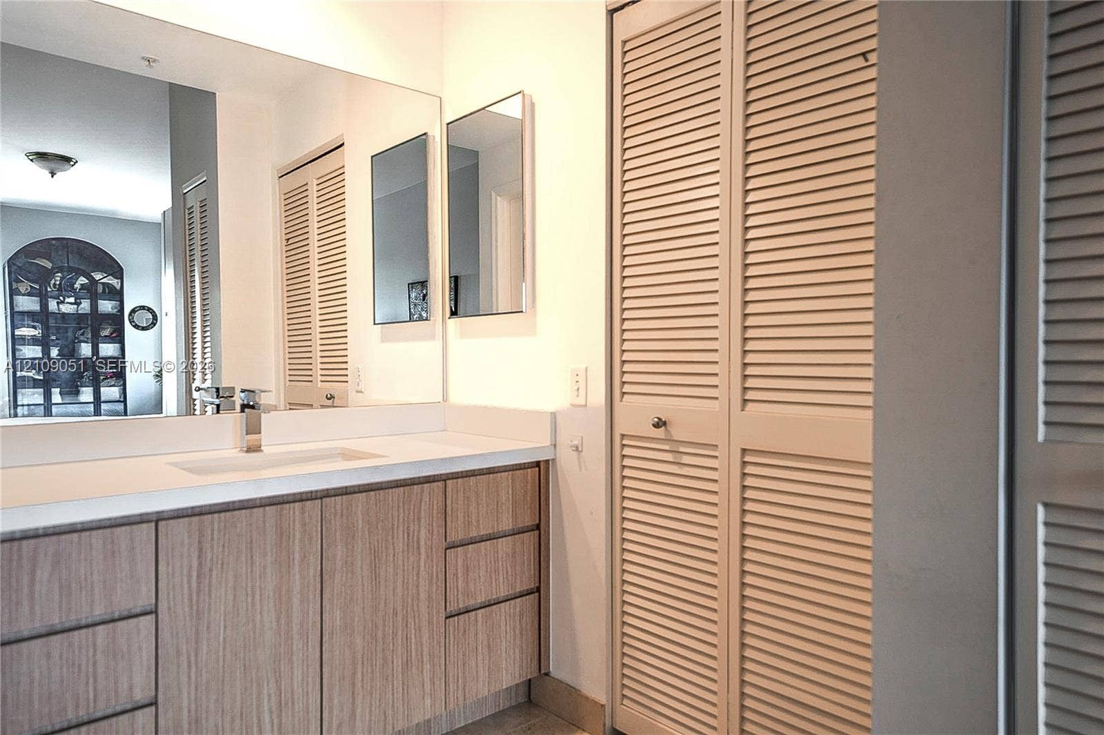 Vanity with wood-finish cabinetry, white countertop, integrated rectangular sink, wall-spanning mirror, and bi-fold louvered closet doors
