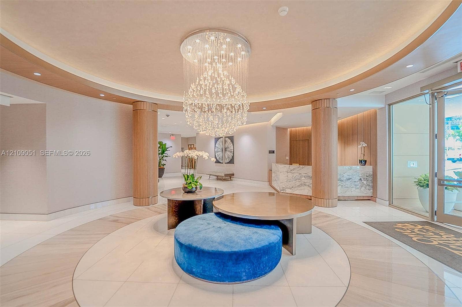 Grand foyer featuring a circular recessed ceiling, integrated crystal chandelier, polished stone flooring, custom wood-paneled pillars, and a stone-clad reception desk