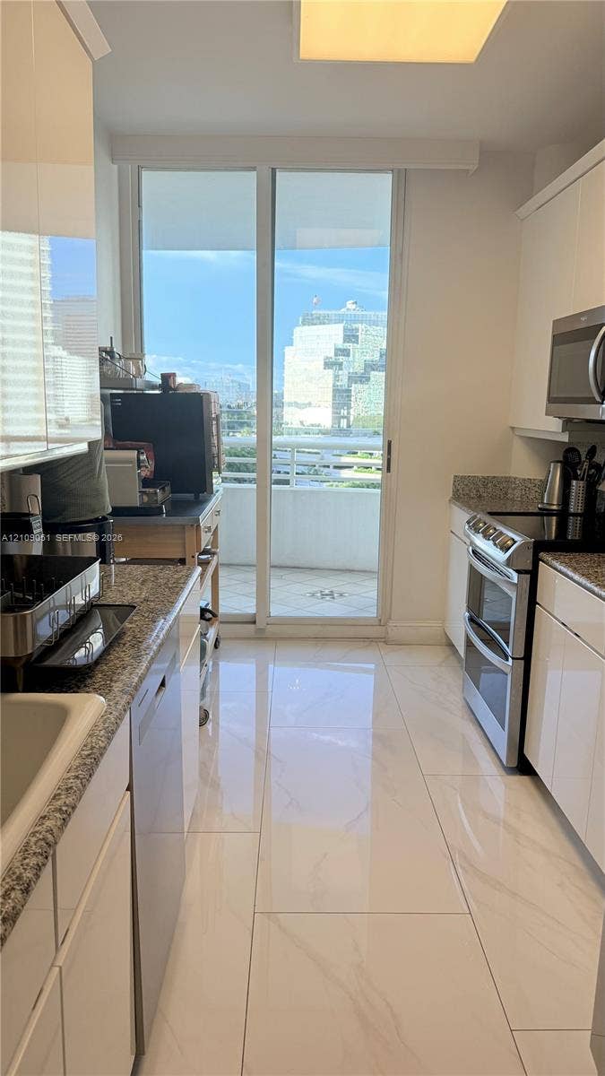 Kitchen featuring high-gloss white cabinetry, stone-finish countertops, stainless steel appliances, large-format tile flooring, and a glass sliding door opening to an exterior balcony