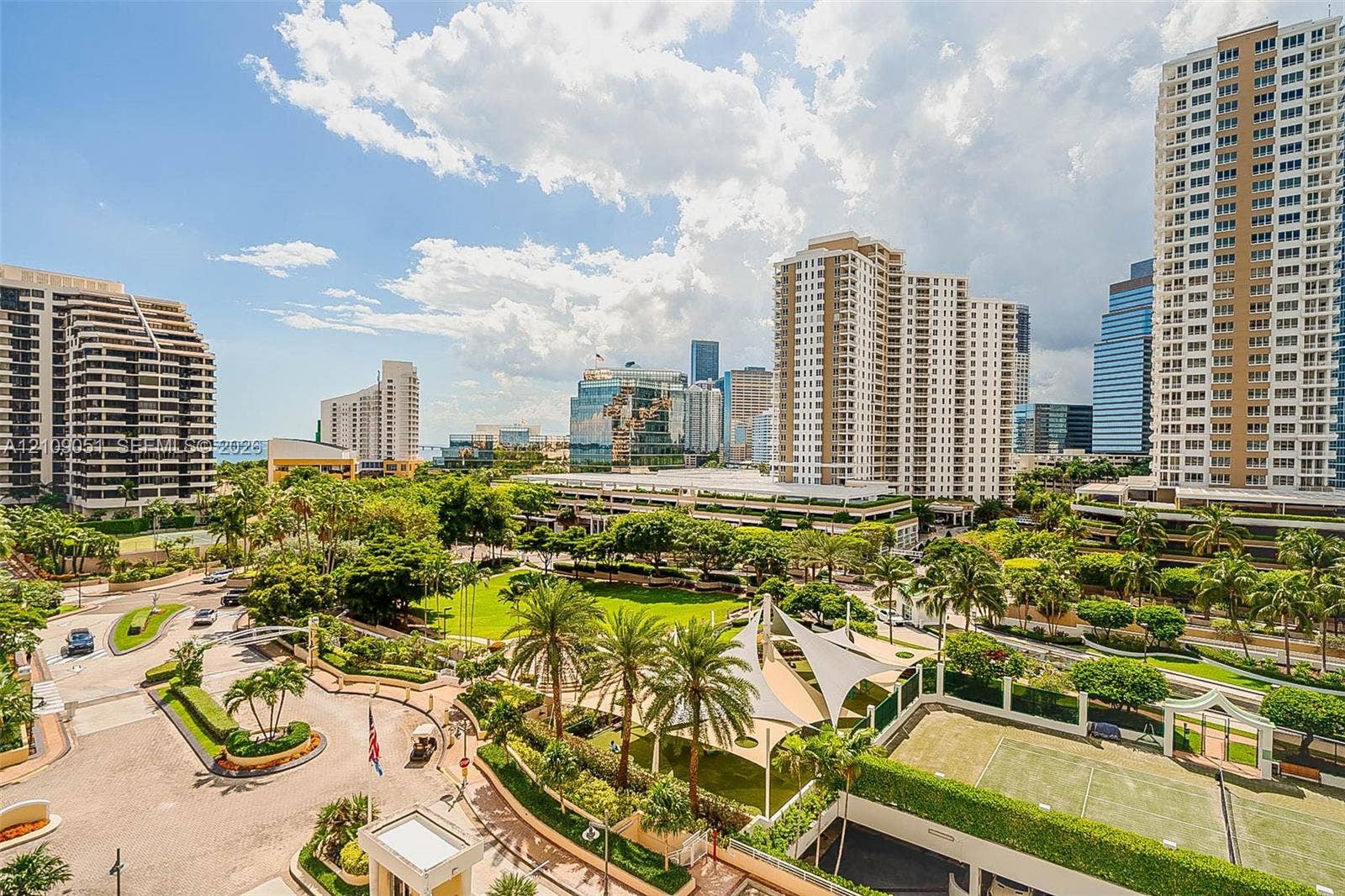 High-rise urban landscape featuring manicured lawns, mature palm trees, tensile shade structures, and an on-site tennis court