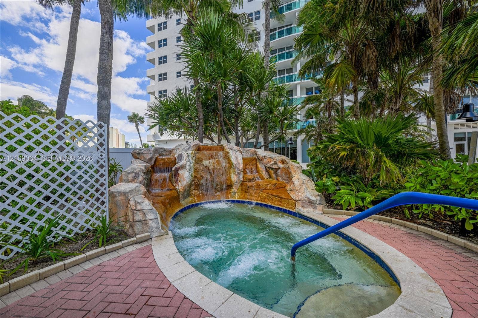 In-ground spa featuring a natural stone waterfall, brick-paver decking, and metal safety handrail