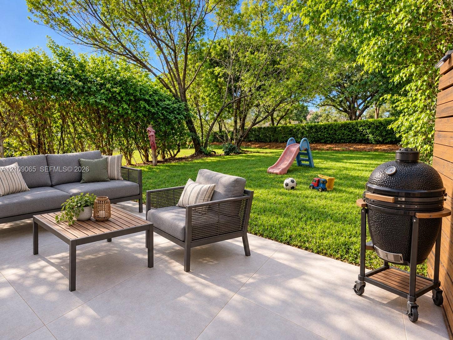 Rear patio featuring large-format light-toned stone tile flooring, horizontal wood-slat privacy fencing, and a grassy lawn bordered by mature trees and manicured hedges