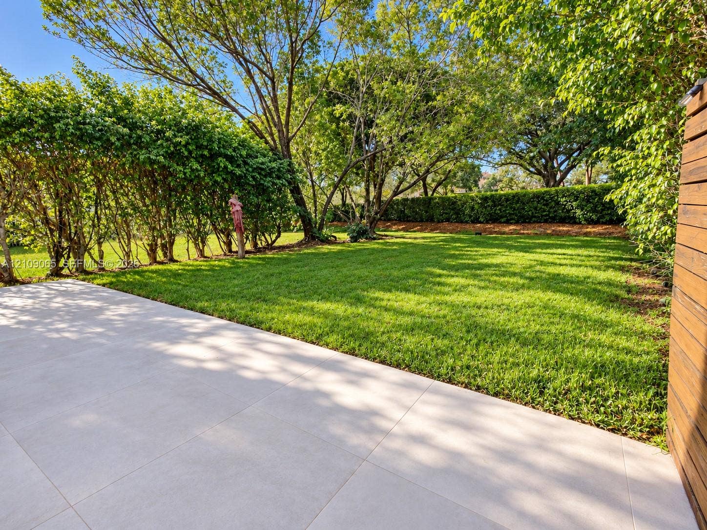 Expansive backyard featuring a smooth-finish paved patio, a sprawling lawn, and mature tree-lined perimeter with established hedges