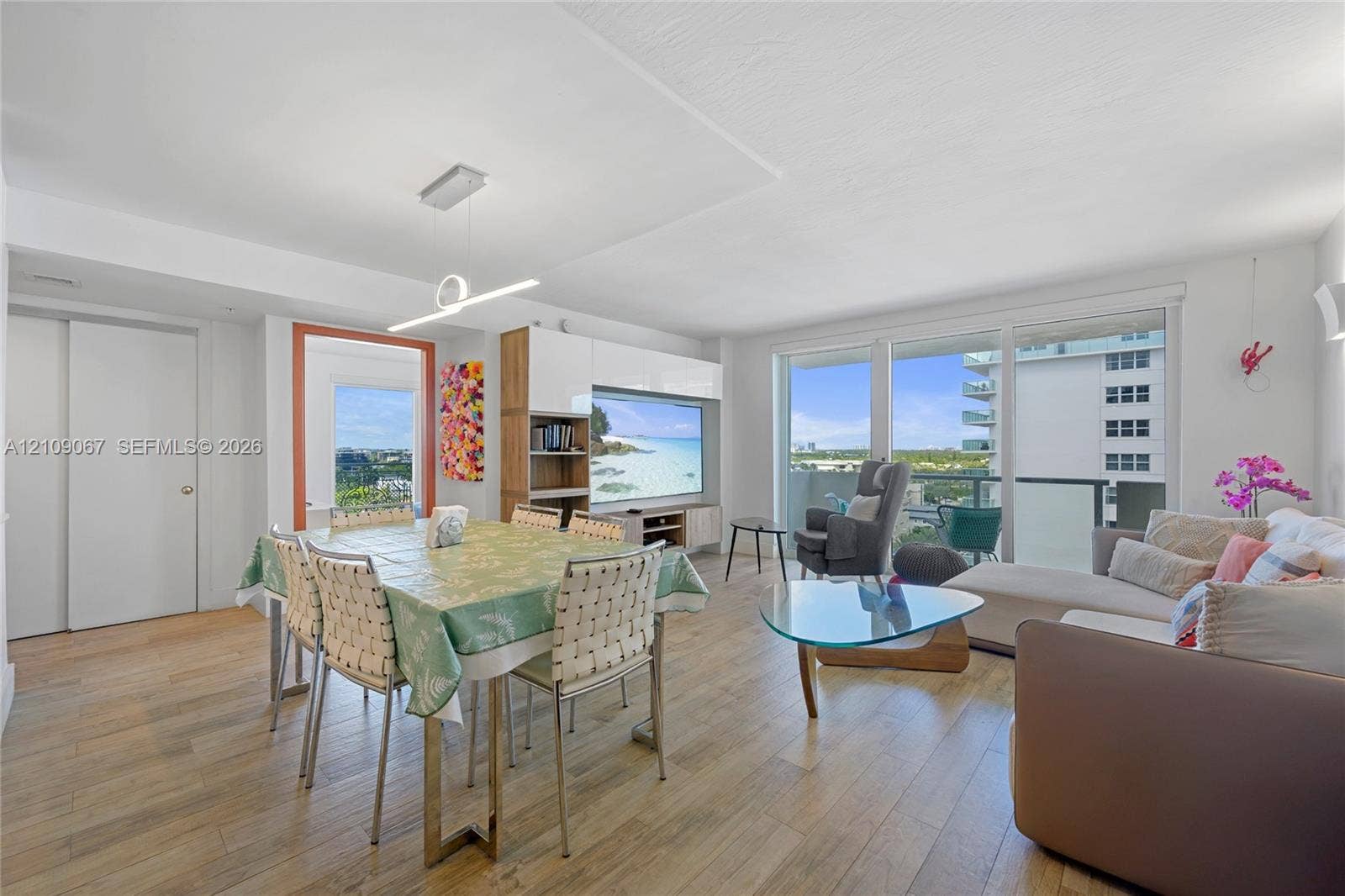 Open-concept living space featuring wood-finish flooring, floor-to-ceiling glass sliding doors, built-in media cabinetry with high-gloss finishes, and contemporary linear pendant lighting