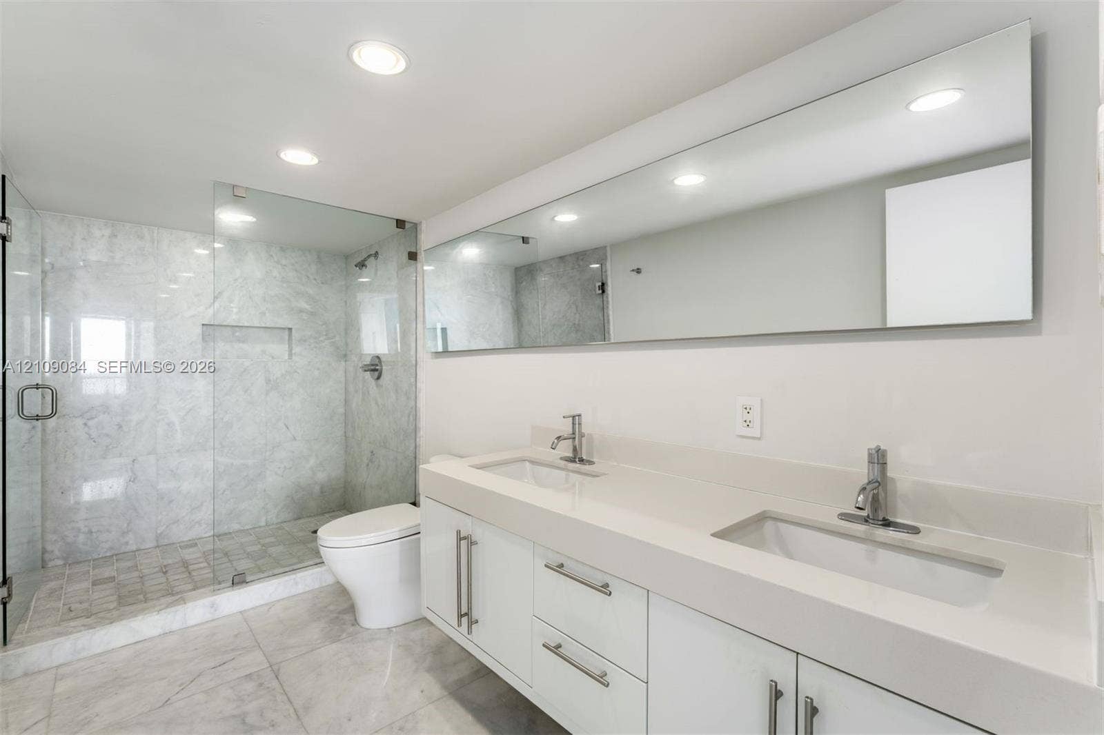 Dual-sink vanity with quartz countertop, walk-in glass-enclosed shower featuring stone-finish tiling, full-width vanity mirror, and recessed lighting