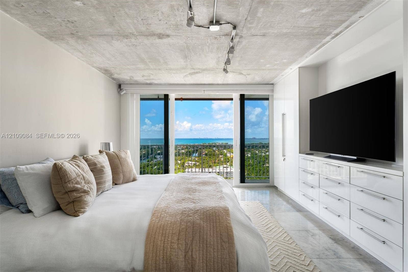 Exposed concrete ceiling with industrial track lighting, floor-to-ceiling glass paneling, built-in high-gloss white cabinetry, polished tile flooring, and panoramic ocean backdrop