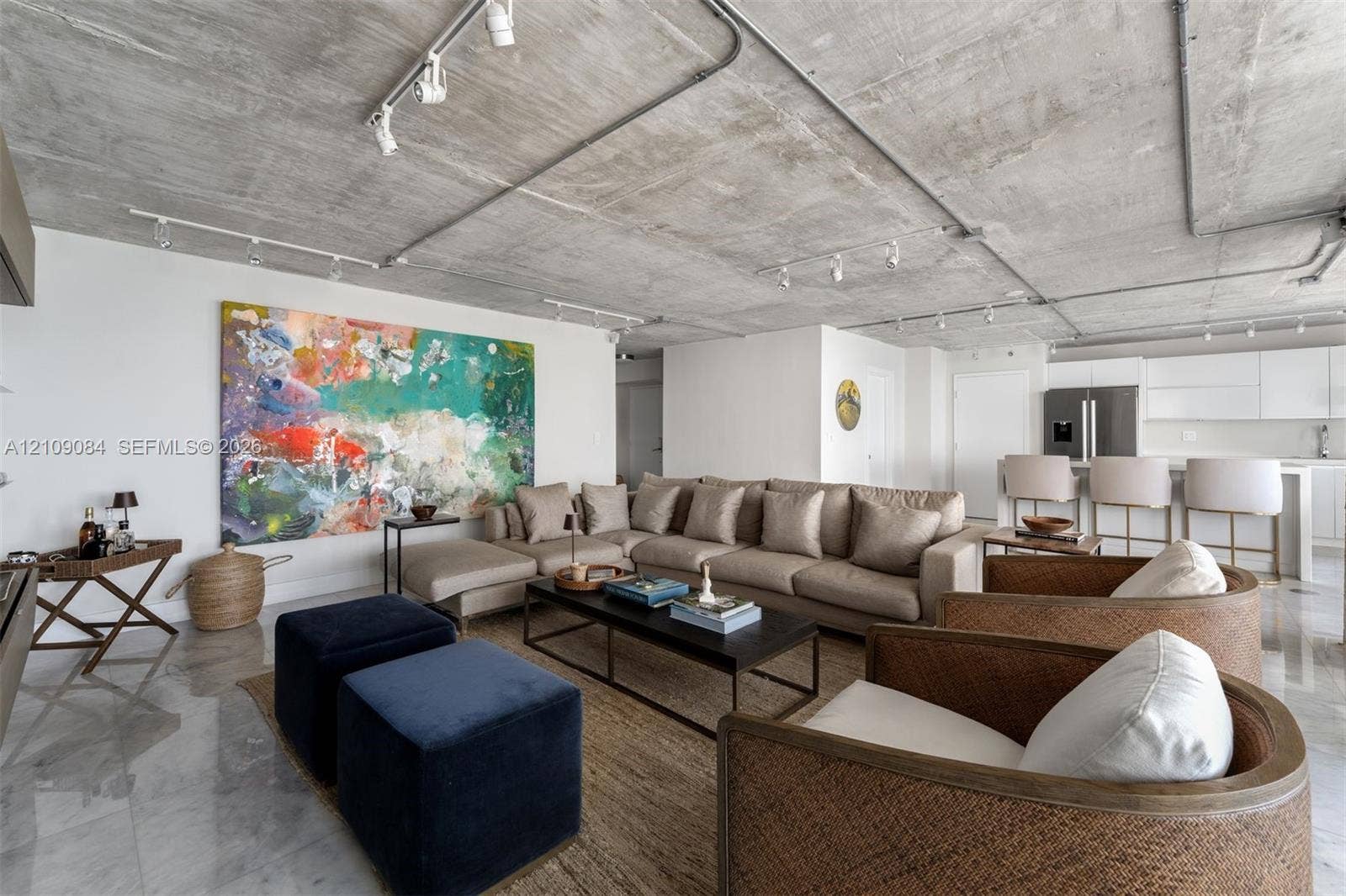 Exposed concrete ceiling with industrial track lighting, floor-to-ceiling glass doors, wall-mounted light wood-finish cabinetry, and balcony access featuring ocean water scenery