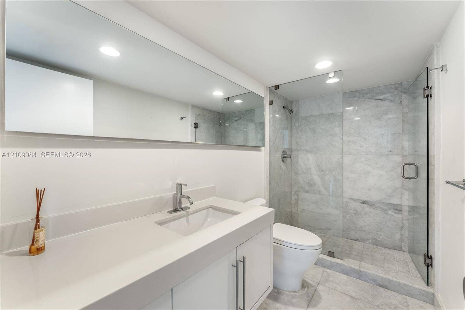 White vanity with a solid-surface countertop and integrated basin, full-width mirror, and walk-in shower featuring glass paneling and stone-tile surrounds