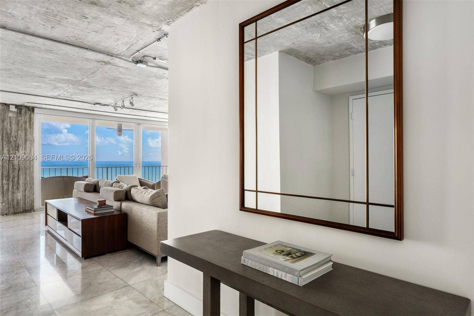 Exposed concrete ceilings, large-format tile flooring, wall-mounted track lighting, and full-height glass doors showcasing ocean scenery