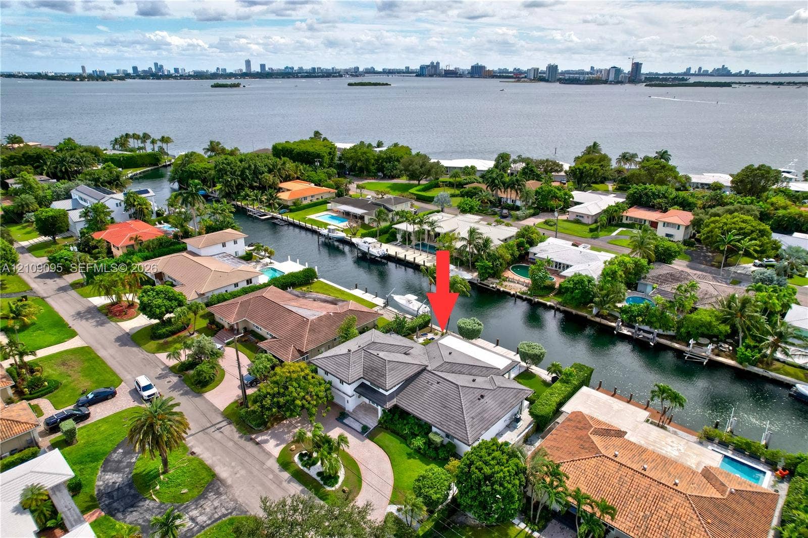 1370 NE 103rd Street, Miami Shores, FL 33138 Waterfront property featuring a canal-side bulkhead, multi-gabled tile roof, paver driveway, and mature landscaping