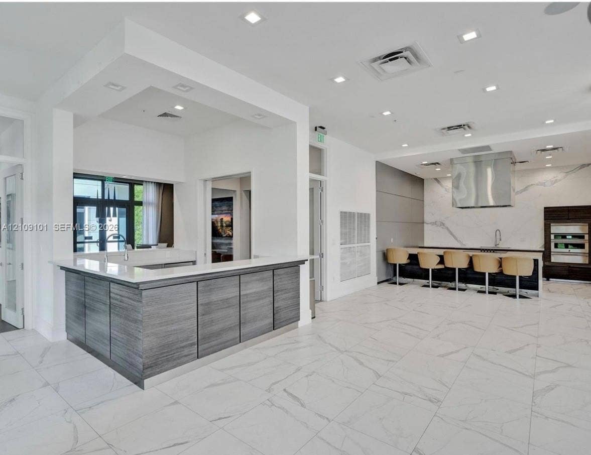 Kitchen featuring large-format tile flooring, a dual-level island with stone countertops and wood-finish cabinetry, a full-height stone backsplash, and stainless steel hood range