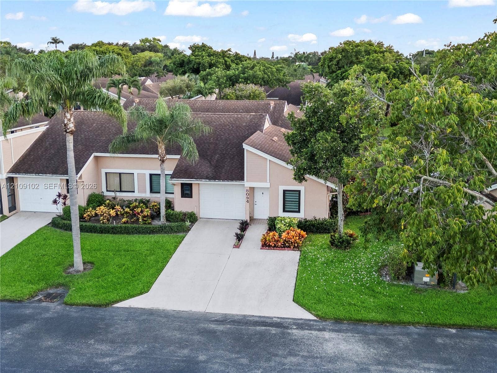 Single-story exterior featuring a shingled roof, concrete driveway, two-car garage, and established tropical landscaping