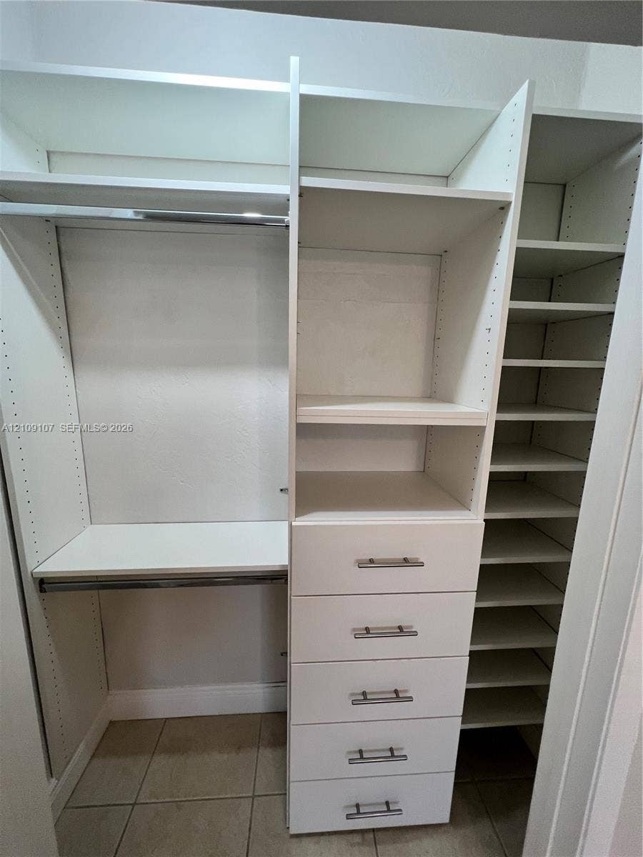 Custom reach-in closet featuring white built-in shelving, a five-drawer stack with satin-nickel hardware, dual clothing rods, and tiled flooring