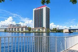Waterfront high-rise building with a white facade and red roofline, featuring balcony-lined exterior walls and a metal railing along the paved bulkhead