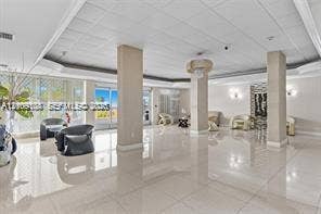 Expansive lobby featuring polished tile flooring, tray ceilings with recessed lighting, structural support columns, wall-mounted accent lighting, and floor-to-ceiling glass paneling