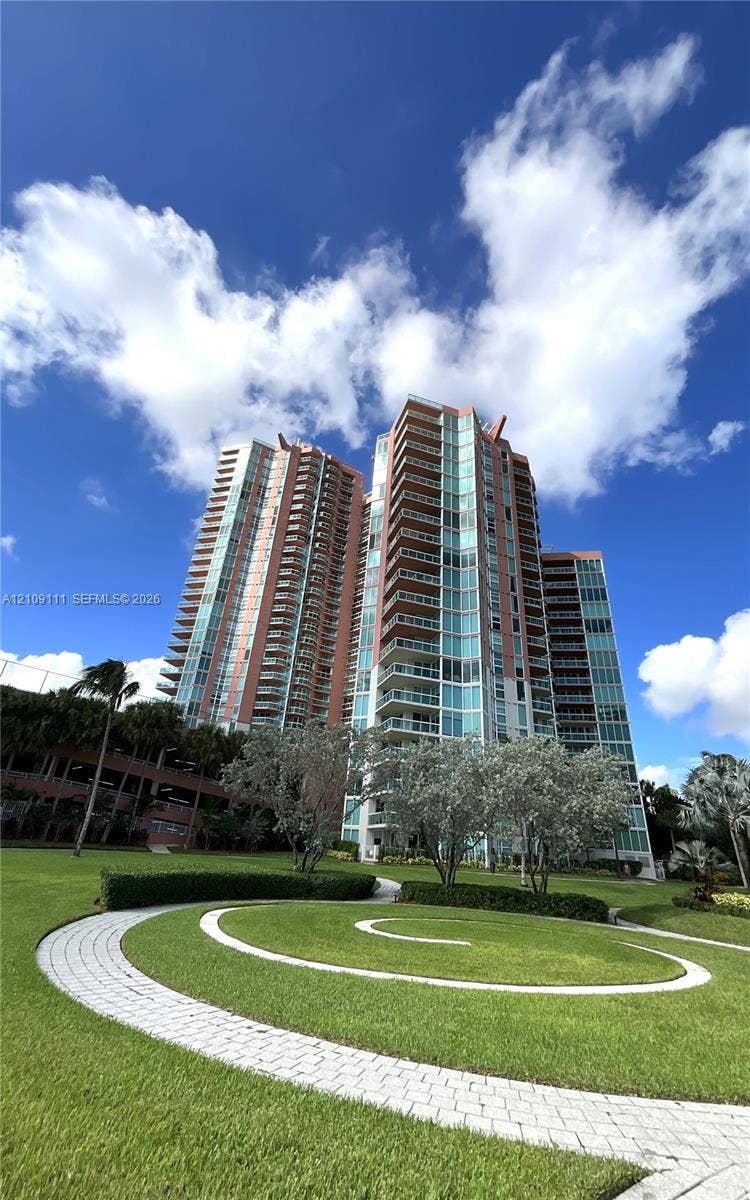 3370 Hidden Bay Drive, #801, Aventura, FL 33180 High-rise residential structure featuring glass facades and wrap-around balconies, complemented by landscaped grounds, manicured lawns, and paved stone walkways