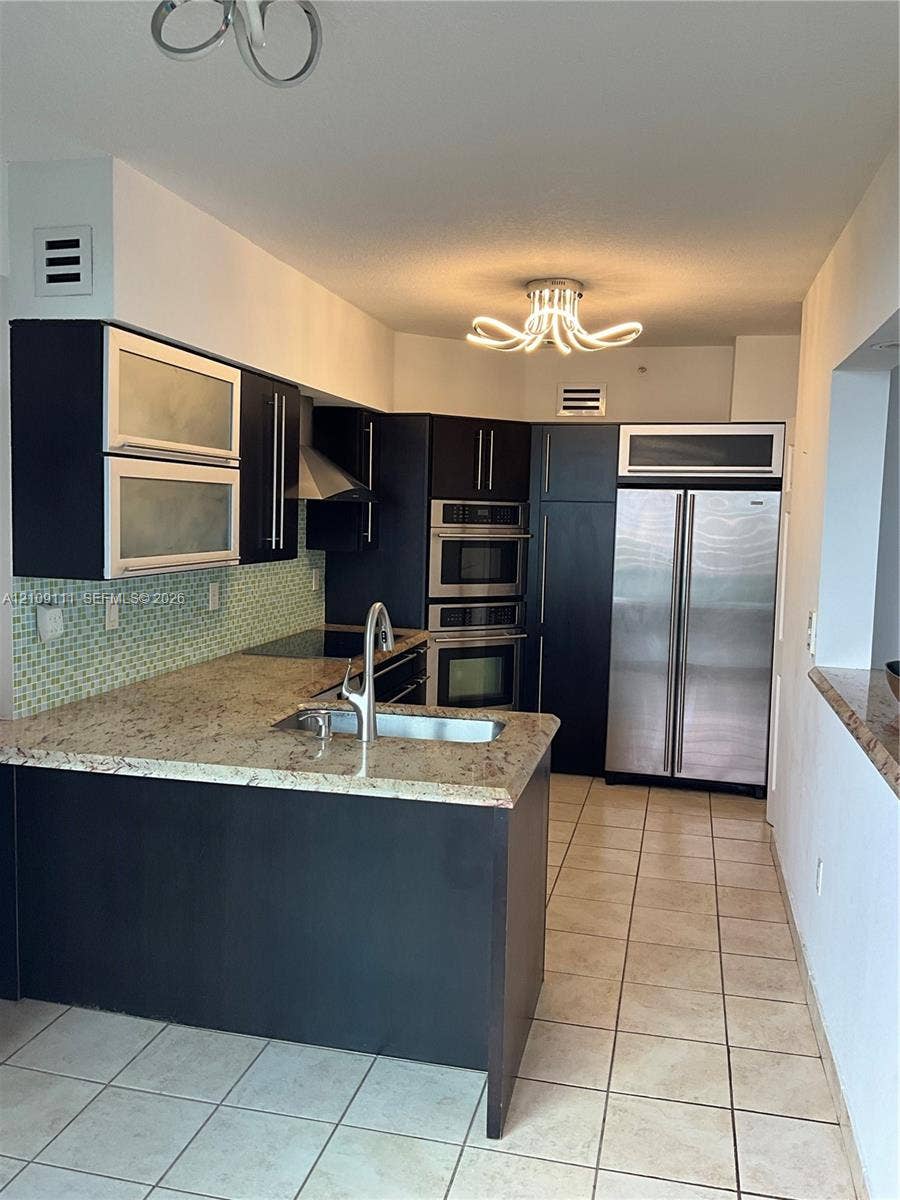 3370 Hidden Bay Drive, #801, Aventura, FL 33180 Kitchen featuring dark cabinetry, stone countertops, a mosaic tile backsplash, integrated stainless steel appliances, and a modern tiered chandelier