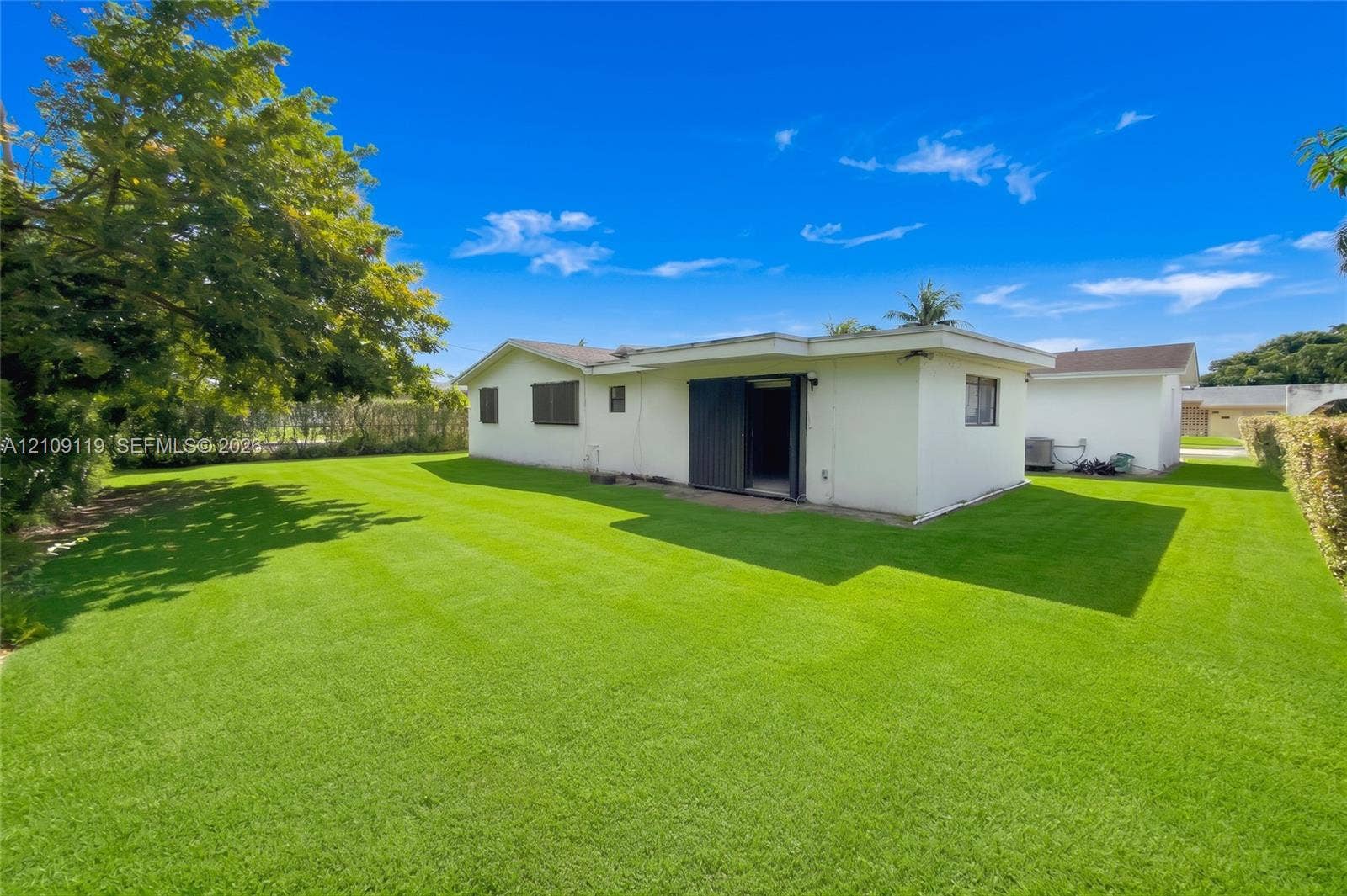 Rear exterior featuring white stucco siding, a flat roofline, sliding glass doors, and an expansive lawn with mature perimeter trees